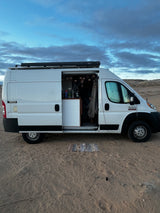 This white RV rental, a spacious camper van with its side door open, reveals a person inside. Parked on sand under cloudy skies.