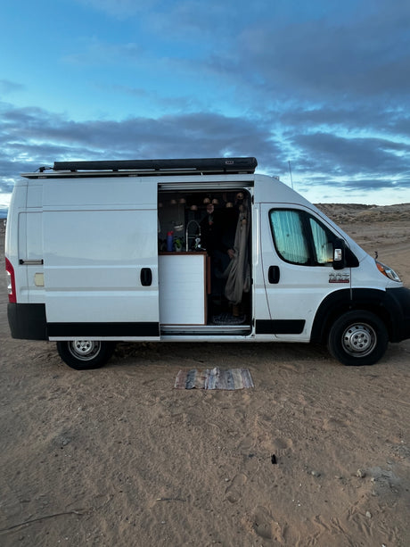 This white RV rental, a spacious camper van with its side door open, reveals a person inside. Parked on sand under cloudy skies.
