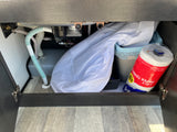 RV rental under-sink cabinet reveals plumbing, a water tank, and practical storage for paper towels & a trash can.