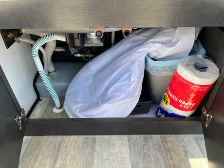 RV rental under-sink cabinet reveals plumbing, a water tank, and practical storage for paper towels & a trash can.