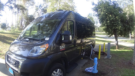 A grey Ram ProMaster camper van, an RV rental, connected to campsite water and sewer hookups on a sunny day.