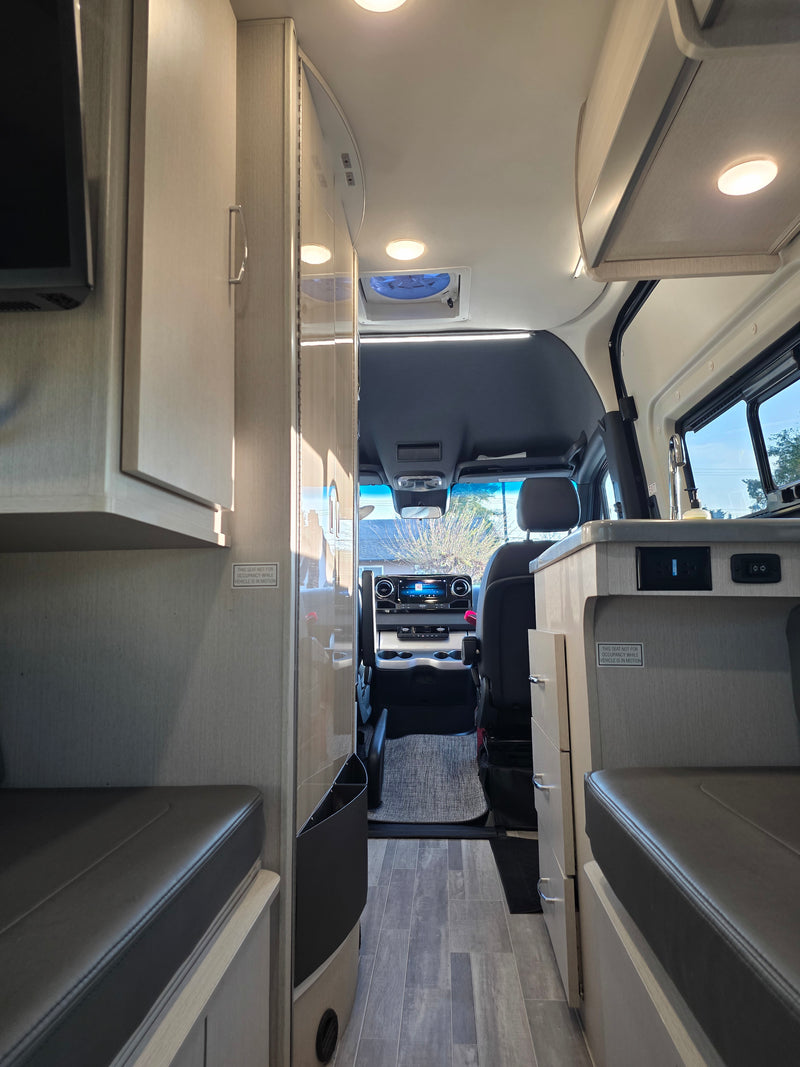 View inside an RV rental showing a narrow aisle leading to the cockpit, flanked by light wood cabinets and gray cushioned benches. — photo 2