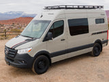 Modern tan Mercedes Sprinter van, an RV rental, with roof rack, parked on dirt with scenic mountains. Perfect camper van for travel.