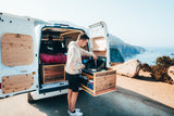 RV rental trip: Man cooks from his camper van's pull-out kitchen, enjoying a stunning ocean view.