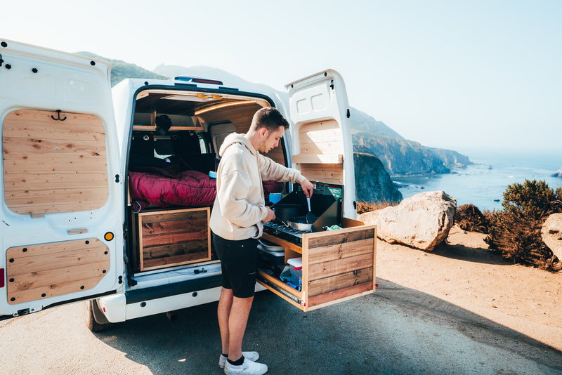  enjoying a stunning ocean view.|Adventure awaits! This custom camper van RV rental offers a comfortable sleeping area & pull-out kitchen with mountain views.|RV rental camper van interior showing a cozy bed and a Coleman propane stove for outdoor cooking at a campsite.|RV rental: Inside a converted camper van — photo 2