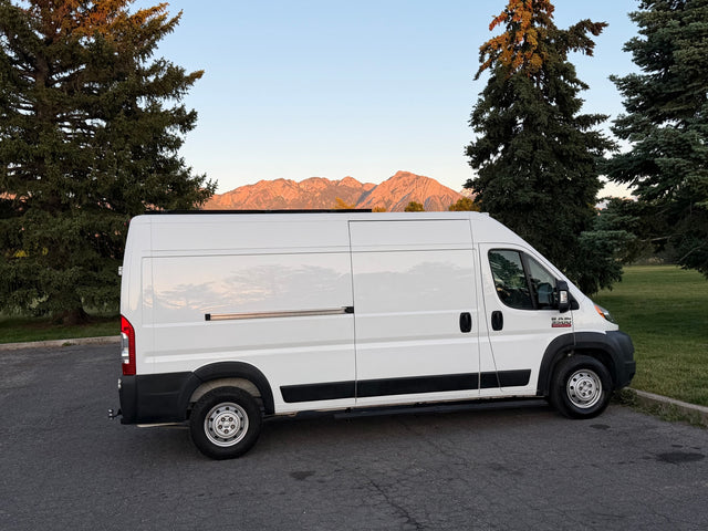 White Ram ProMaster camper van (RV rental) parked on asphalt, with sunlit mountains and trees under a clear sky.