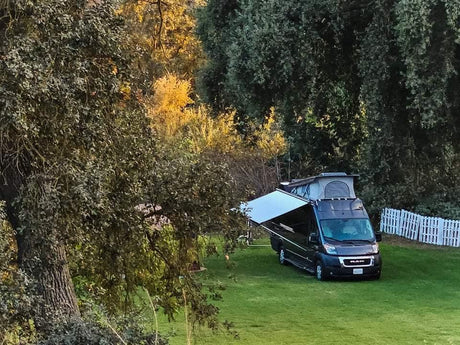 This RV rental with a pop-top and awning sits on a green lawn, surrounded by lush trees and a white fence.