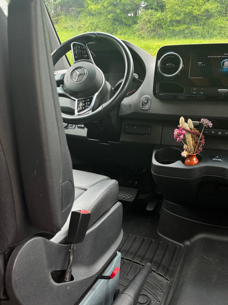 Inside an RV rental, showing the Mercedes-Benz steering wheel, dashboard, and a vase of dried flowers near the infotainment screen.