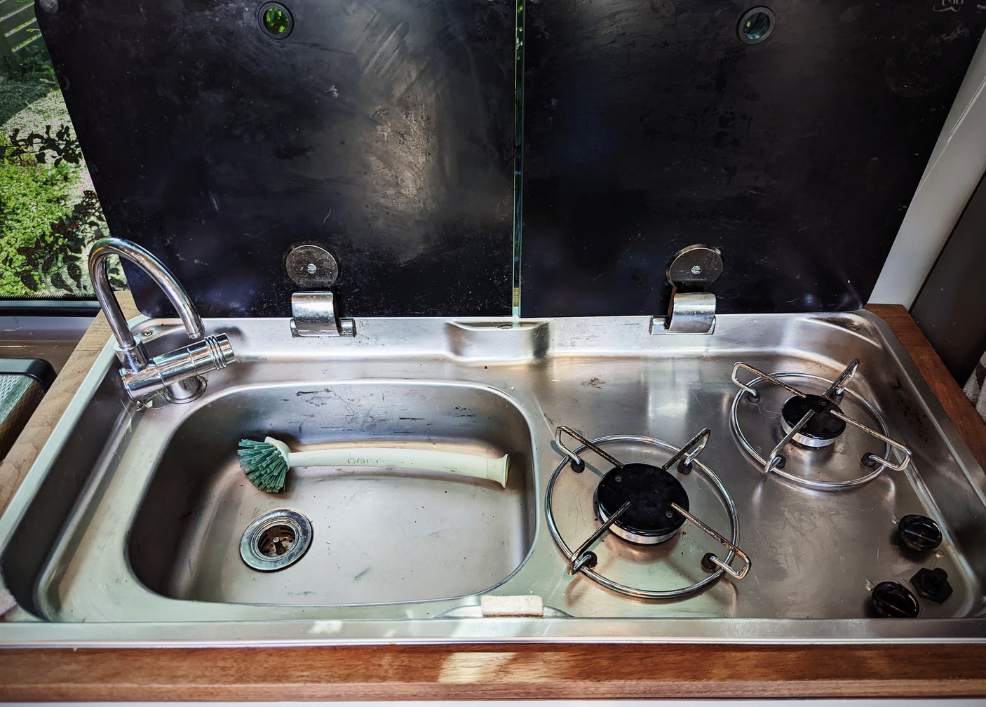 Open black covers reveal a compact RV rental kitchen: stainless steel sink, faucet, and two-burner stove. Ready for meal prep on the go!
