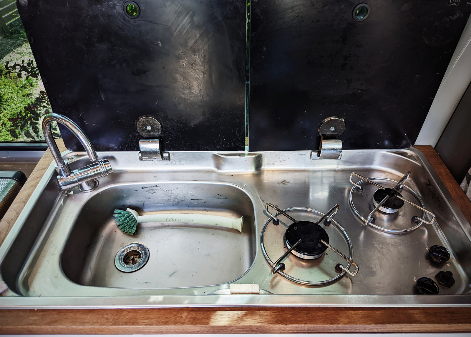 Open black covers reveal a compact RV rental kitchen: stainless steel sink, faucet, and two-burner stove. Ready for meal prep on the go!