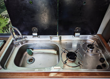 Open black covers reveal a compact RV rental kitchen: stainless steel sink, faucet, and two-burner stove. Ready for meal prep on the go!