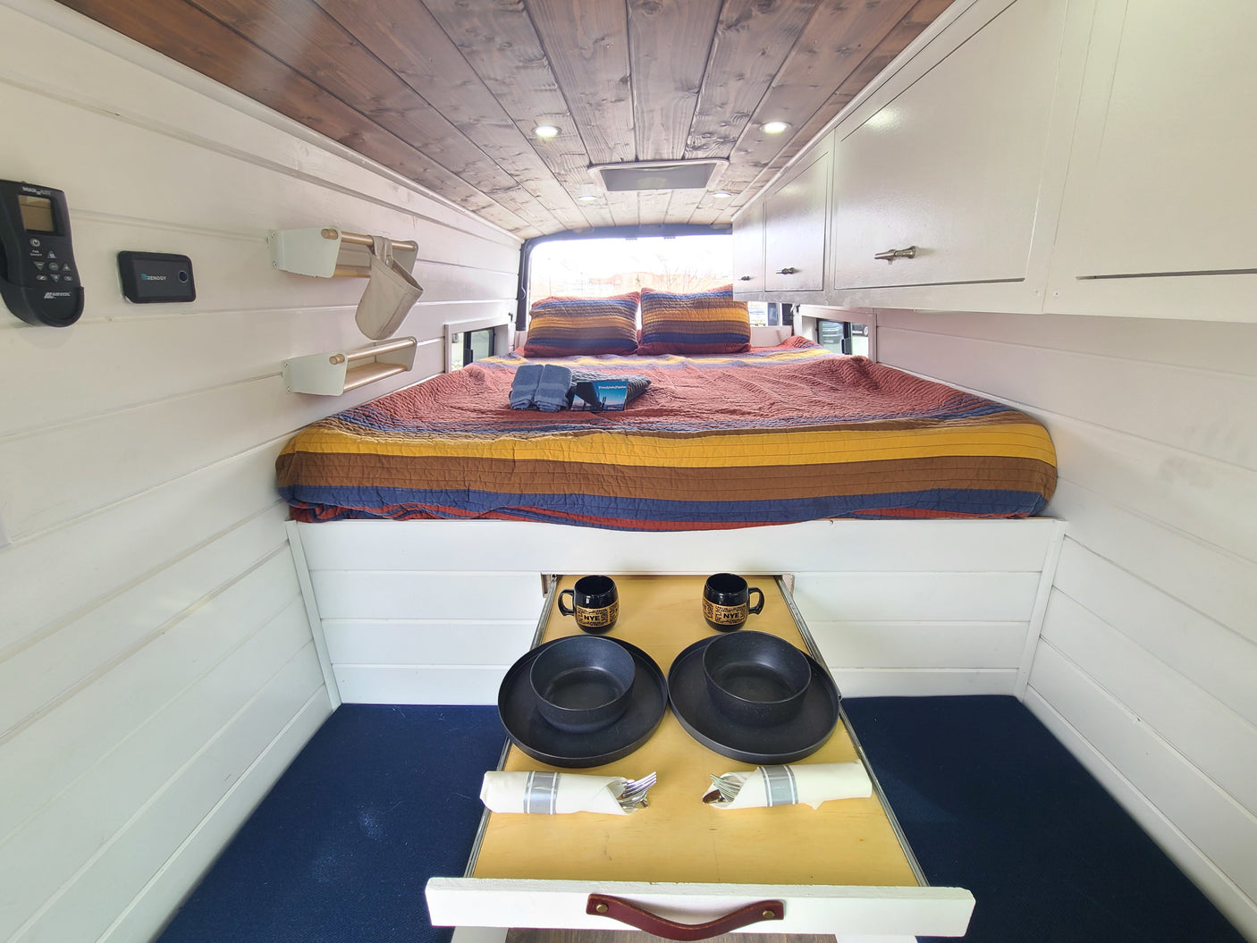 Cozy RV rental interior: elevated bed with colorful quilt, white shiplap walls, wood ceiling, & pull-out dining table.