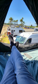Feet up in an RV rental! View shows parked campervans, palm trees, and a sunny beach scene, offering relaxing travel vibes.