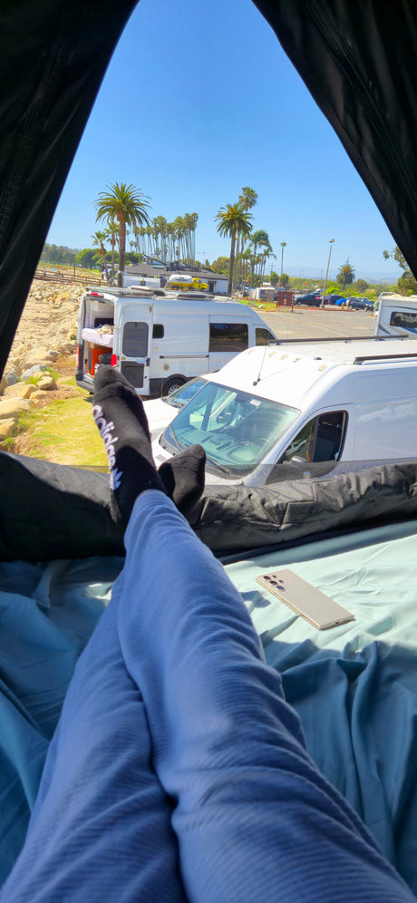 Feet up in an RV rental! View shows parked campervans, palm trees, and a sunny beach scene, offering relaxing travel vibes.
