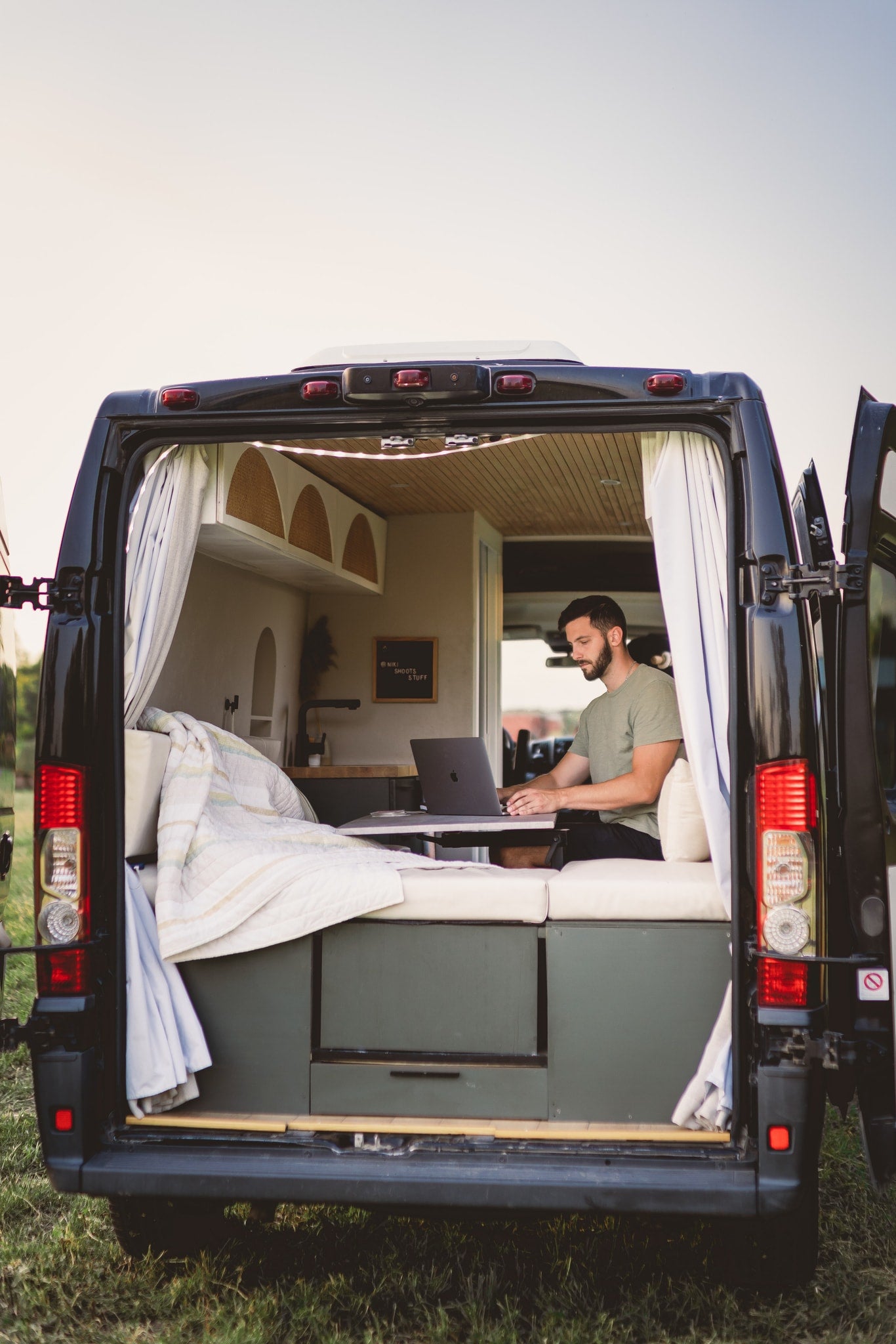 Man on laptop inside a black, converted RV rental van with rear doors open. Perfect for remote work and digital nomad adventures. — photo 10