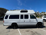 White high-top camper van, ideal for rv rental, featuring tinted windows and rugged black wheels, parked under a clear sky.