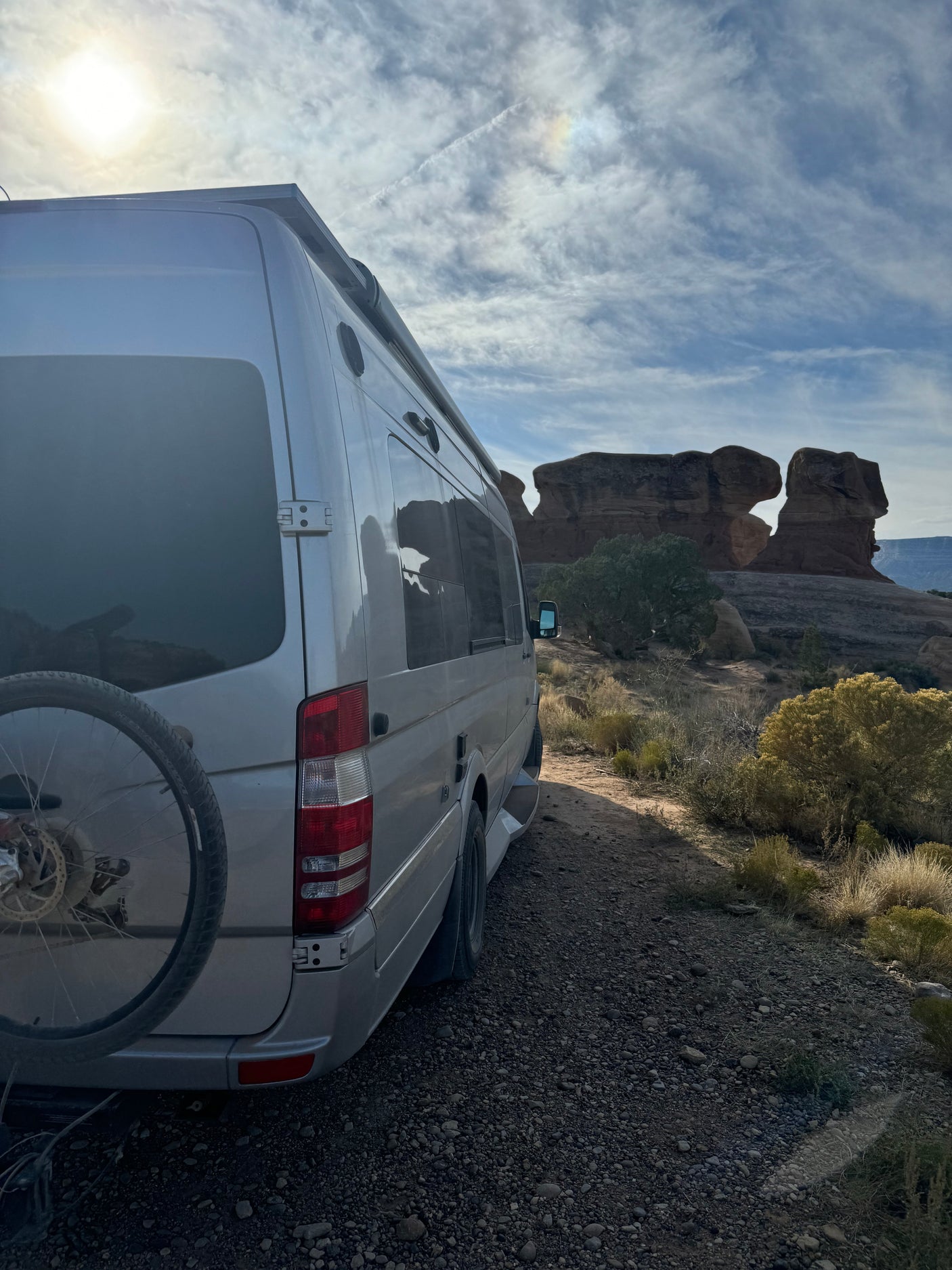 An RV rental camper van with a bike rack is parked on a dirt road, facing unique desert rock formations under a bright sky.