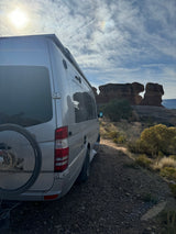 An RV rental camper van with a bike rack is parked on a dirt road, facing unique desert rock formations under a bright sky.