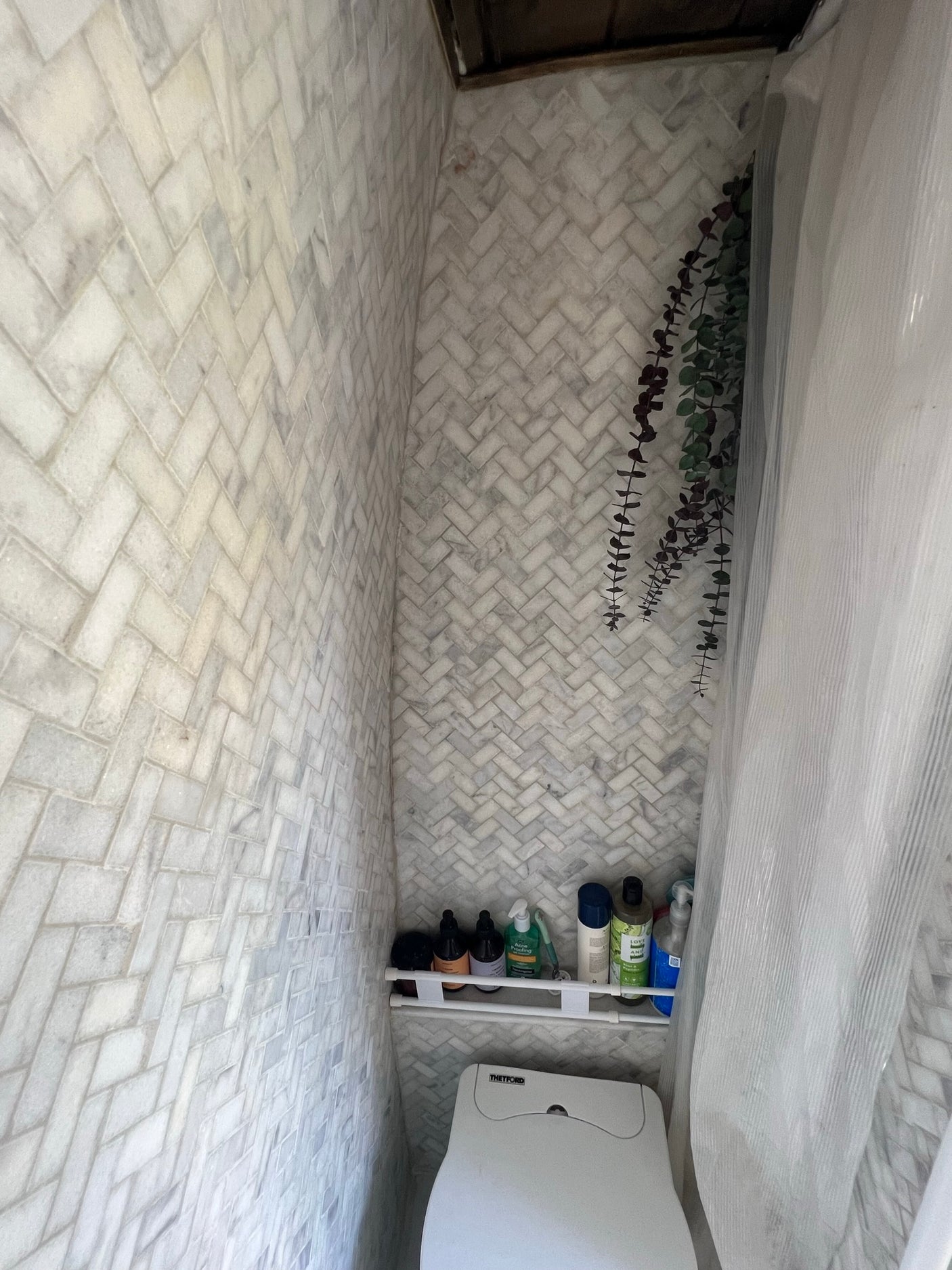 An RV rental's compact bathroom: white herringbone tile walls, toilet, shelf of toiletries, and a shower curtain.