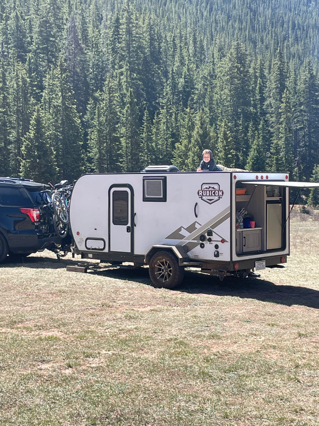 Happy child on a white travel trailer (RV rental) hitched to an SUV with bikes, set against a forest backdrop.