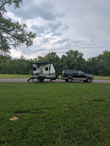 No Boundaries RV rental trailer hitched to a black SUV, set up for camping on a grassy site under cloudy skies.