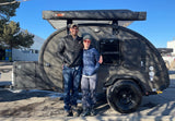 A happy couple poses with their sleek, dark grey teardrop camper, ideal for an RV rental adventure. Equipped with a 23ZERO awning.
