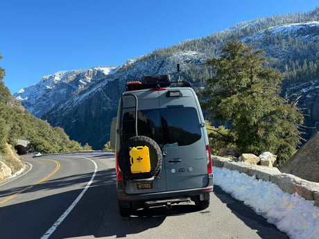 Explore winter wonders in a gray rv rental van on a snowy mountain road, flanked by majestic peaks & a tunnel ahead.