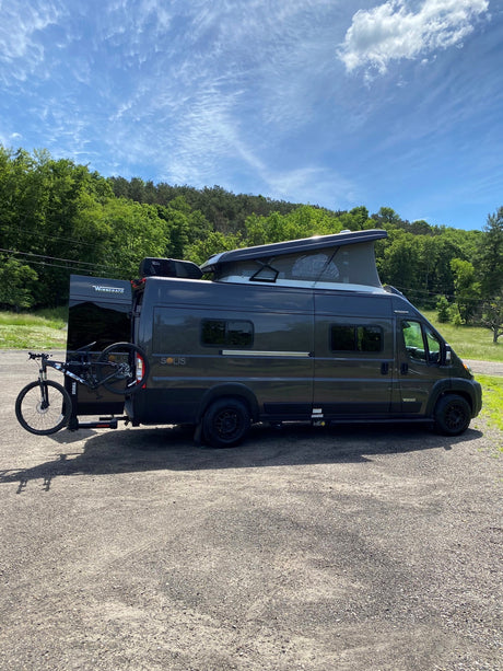 Winnebago Solis RV rental ready for adventure: dark gray van with pop-top, bike rack, parked amidst trees.
