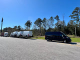 A black van (potential RV rental) parked beside a truck hauling new white vans under a clear sky.