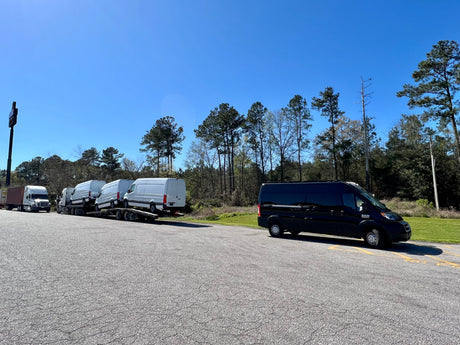 A black van (potential RV rental) parked beside a truck hauling new white vans under a clear sky.