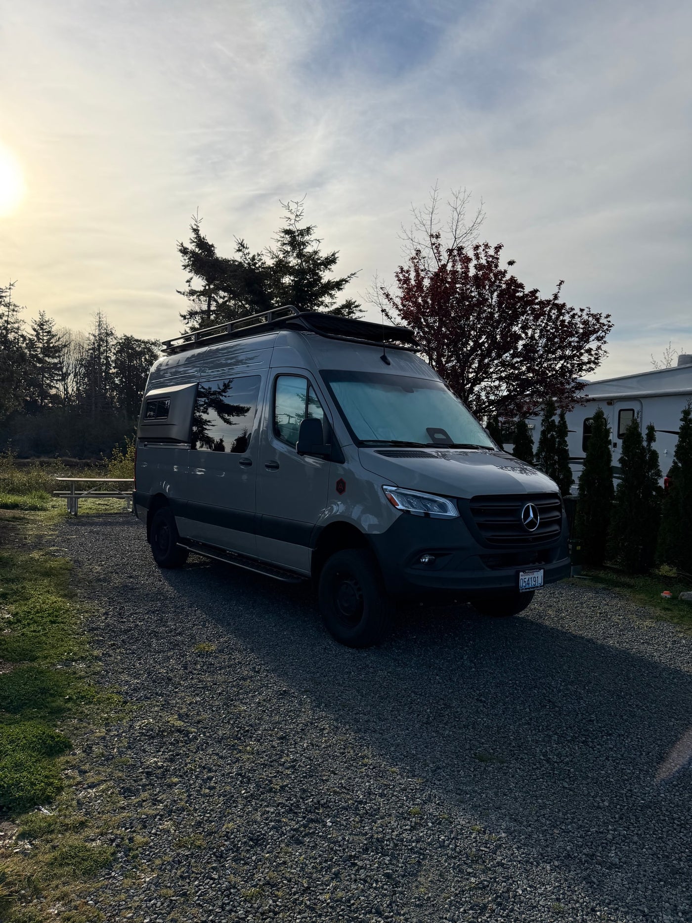 Sunny view of a gray Mercedes Sprinter camper van rv rental at a gravel campsite, trees and another RV nearby. — photo 11