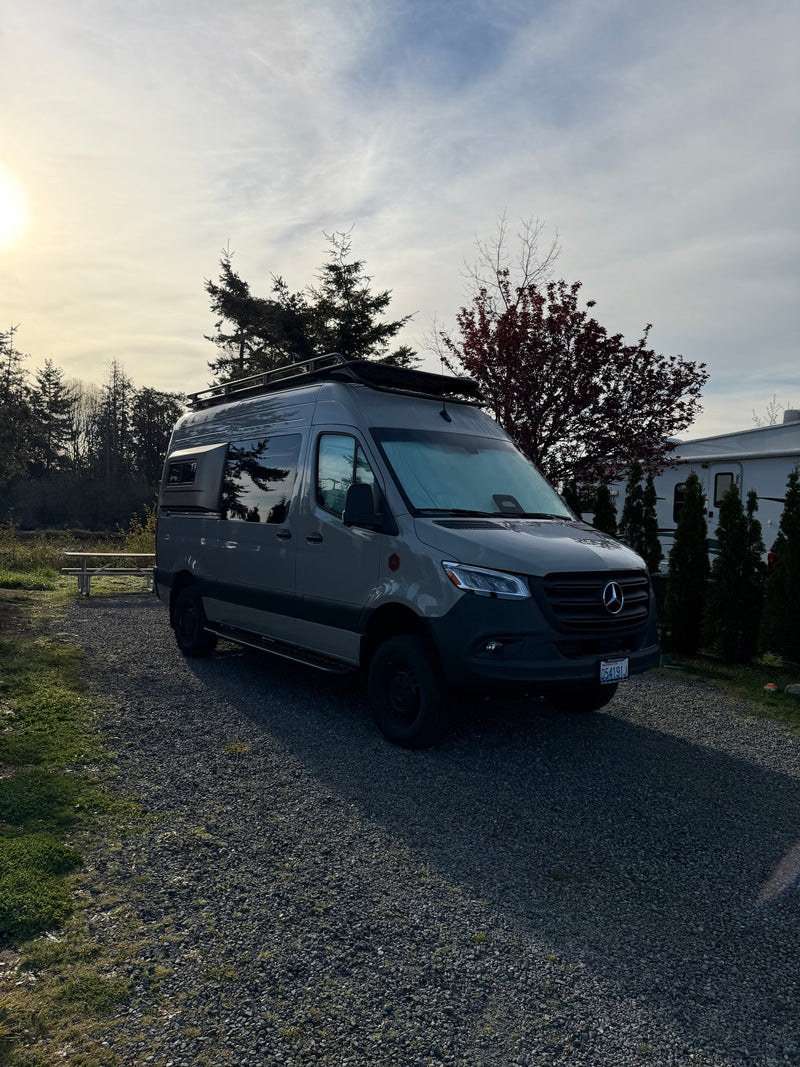 Sunny view of a gray Mercedes Sprinter camper van rv rental at a gravel campsite, trees and another RV nearby. — photo 11