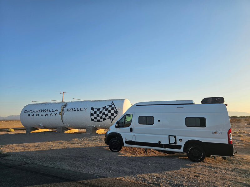 is parked in a golden desert landscape with distant mountains under a vast blue sky.|White RV rental camper van parked by a large Chuckwalla Valley Raceway tank in a sunny desert landscape. Mountains visible.|Step inside this modern rv rental — photo 7