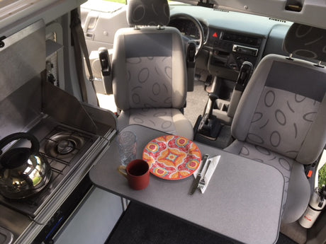 Inside a compact RV rental, showing a kitchen with stove, and a table set with dishes & seating.