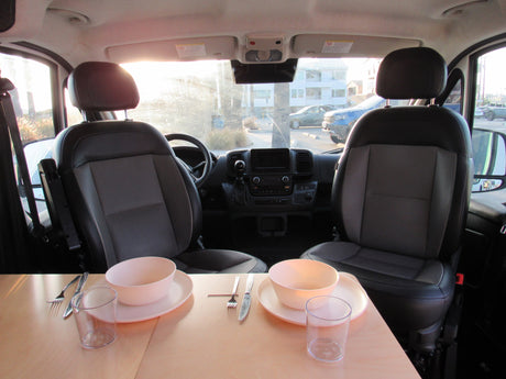 Cozy interior of an RV rental camper van with front seats and a dining table set for two. Bright view out the windshield.