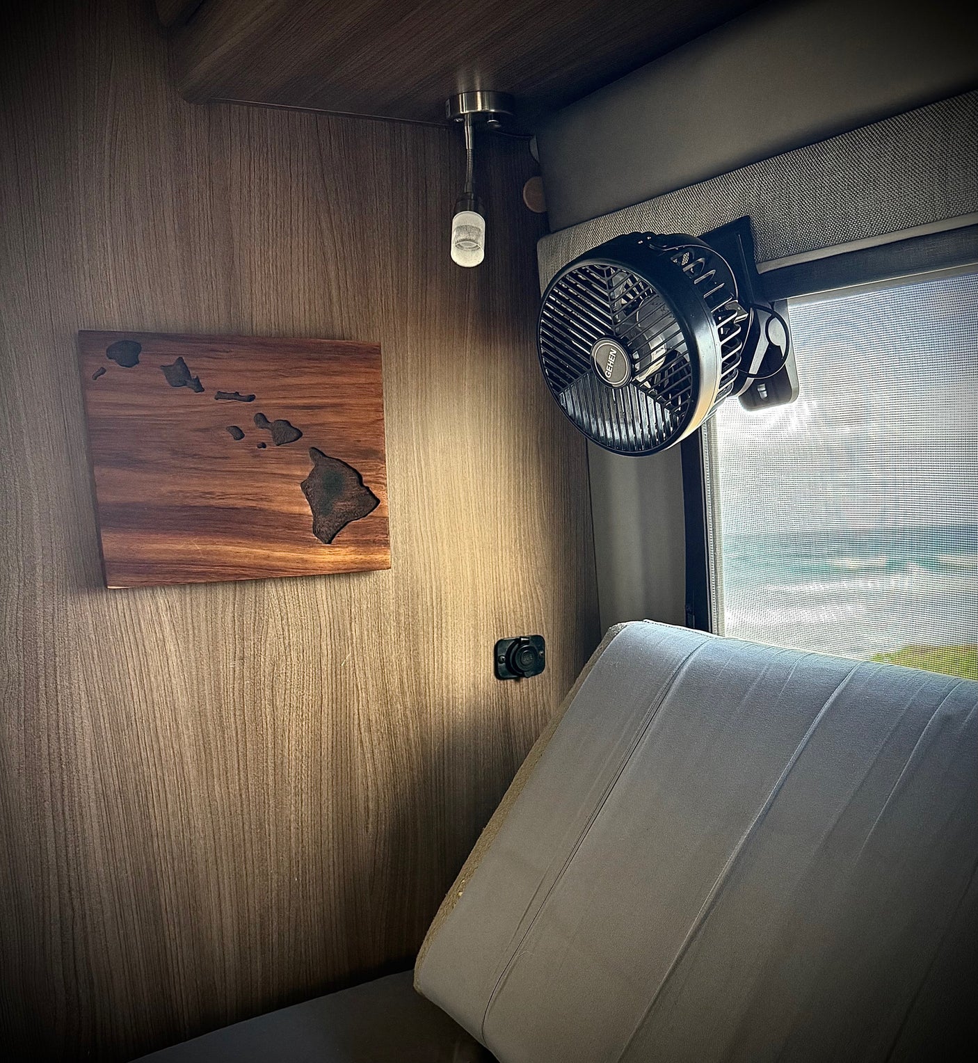 Cozy RV rental interior with Hawaiian map art, a fan, charging port, and an inviting ocean view through the window.