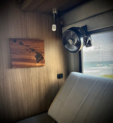 Cozy RV rental interior with Hawaiian map art, a fan, charging port, and an inviting ocean view through the window.