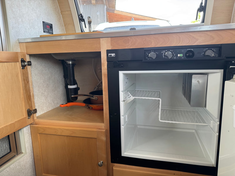 An RV rental's compact galley features an open Norcold refrigerator, sink, and water tank monitor. Pans are under the sink. — photo 16