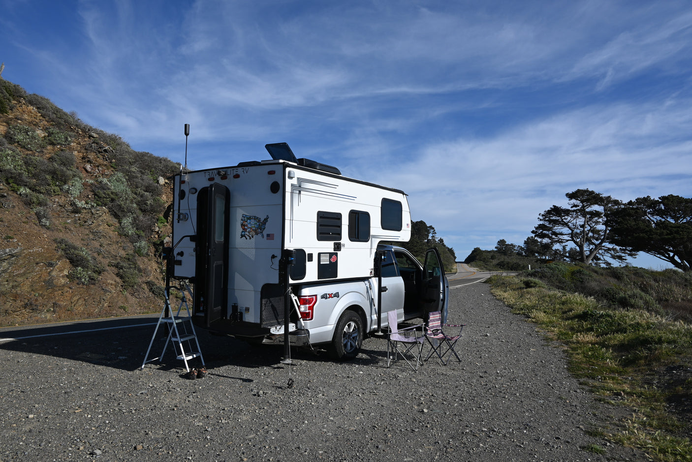 White truck camper RV rental parked roadside with chairs, ladder, and scenic views.