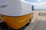 This vibrant yellow and white Scamp travel trailer, ideal for an rv rental, rests by a sunny beach and ocean.