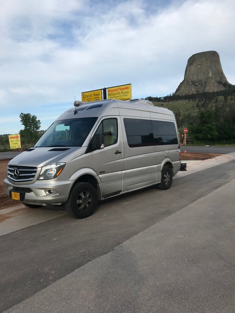 Silver Mercedes Sprinter RV rental van parked with Devils Tower in the background, near signs for KOA amenities.