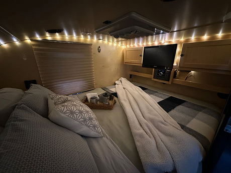 A cozy RV rental bedroom. Bed with patterned pillows, string lights, TV, and soft blanket create a warm, inviting space.