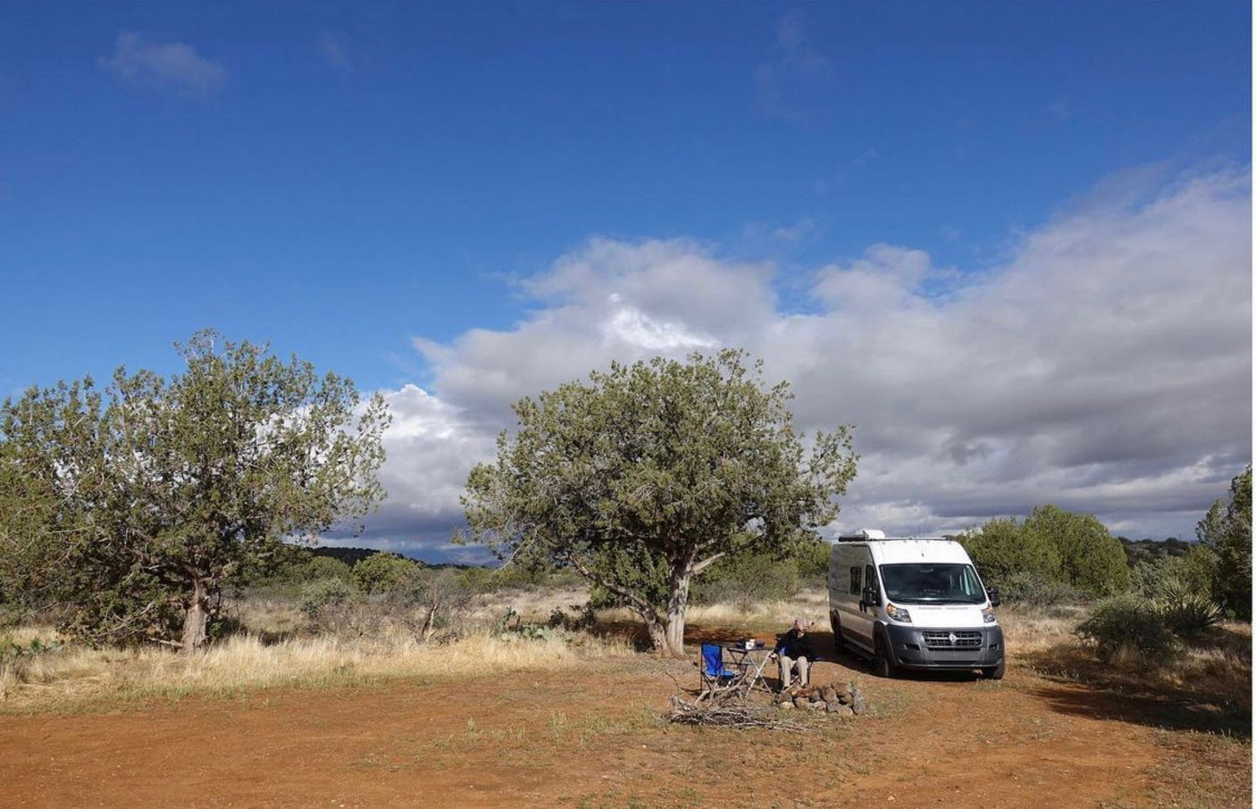 An RV rental camper van parked in a scenic outdoor setting with a person relaxing by a fire pit.