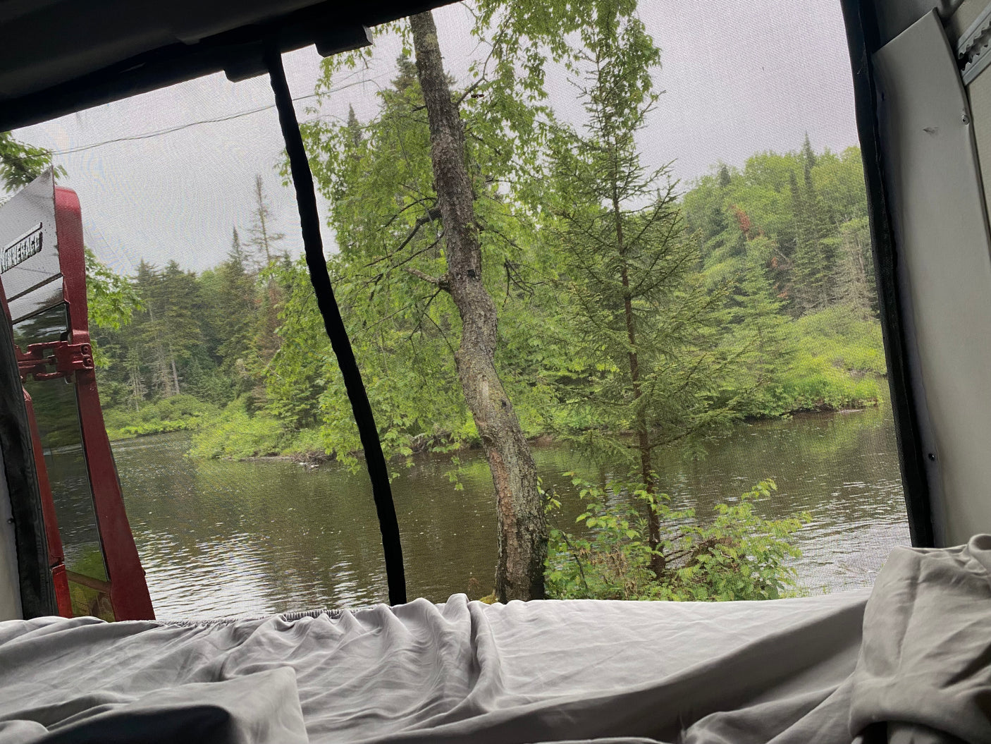 Enjoy a serene RV rental view from bed, featuring a screen window overlooking a tranquil lake and lush forest.