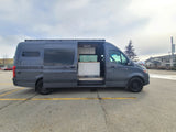A dark grey RV rental van is parked with its side door open, revealing a clean, custom-built interior with white cabinets.