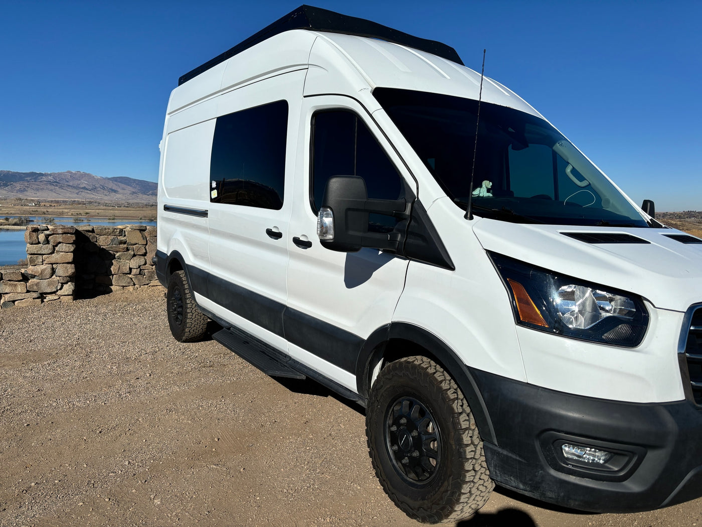 White Ford Transit camper van, an RV rental, on a gravel road with mountains and a lake in the background. — photo 3