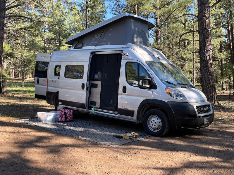 Winnebago Solis rv rental with pop-top open, parked on a dirt campsite among pine trees. Outdoor mat and pet bowls visible. — photo 6