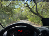 Driving an RV rental through a winding, tree-lined forest road. The dashboard shows 295 miles of range, viewed from the driver's seat.