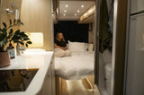 Modern RV rental interior with a woman on the bed, showing kitchen, living, and bathroom areas.