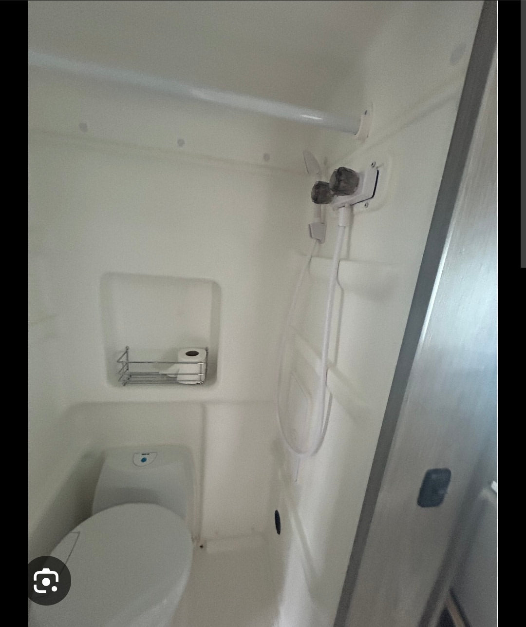 Compact white RV rental wet bath featuring a toilet, shower controls with hose, and a small shelf holding toilet paper.